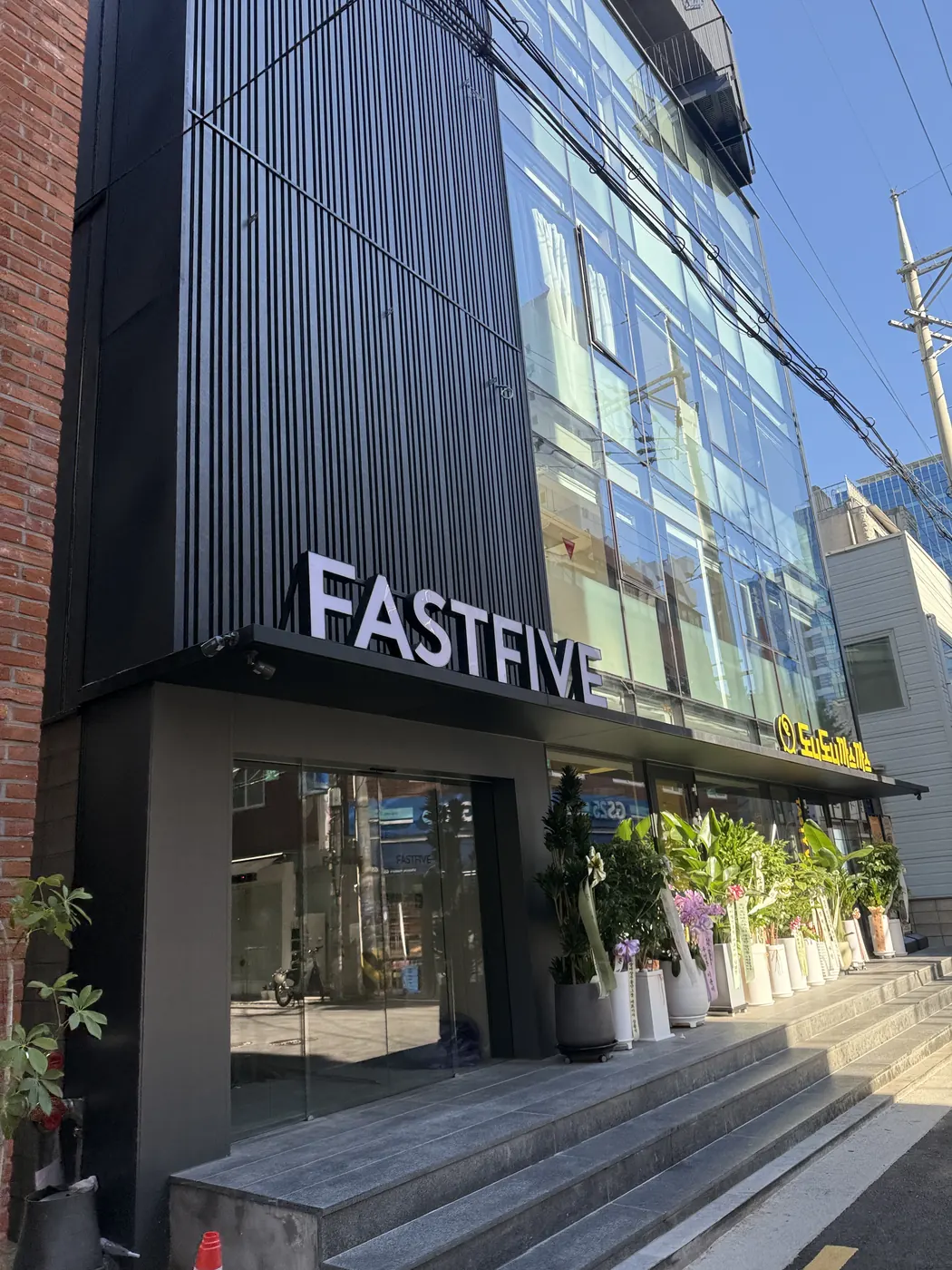 검은 세로 루버 외벽에 FASTFIVE 간판이 붙은 뚝섬점 건물 입구. 계단 옆에 화분이 줄지어 있다